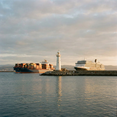 Nautical wall decor: a cruise ship and a container ship passing a white lighthouse at a harbor mouth in late afternoon light