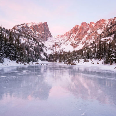 Winter wall art, a frozen mountain lake at sunrise with snowy pines on the shore and pink light on the peaks
